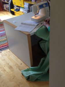Kuvassa pöydänkulma ja osa ompelukonetta, lattialla mintunvihreää kangasta.